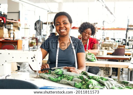 Two black African Zimbabwean ladies working in a textile factory on the sewing line using sewing machines, measuring tapes and scissors to stitch lining in African styled patterned fabric garment Royalty-Free Stock Photo #2011811573