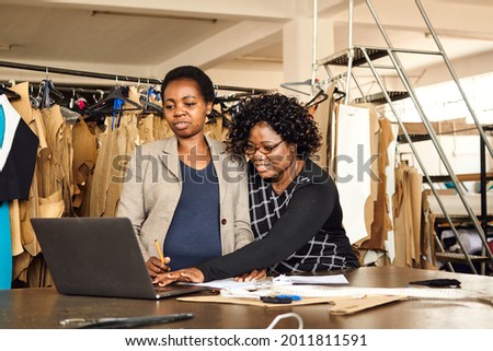 A black African Zimbabwean woman working with colleague processing paperwork with a laptop in textile factory with clothing patterns in the background wearing corporate wear Royalty-Free Stock Photo #2011811591