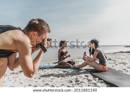 Close up shot of photographer taking picture group of woman getting rest after fitness exercises on the beach. Vlogging, blogging concept. Photoshot of young athletes, sportswomen outdoors.
