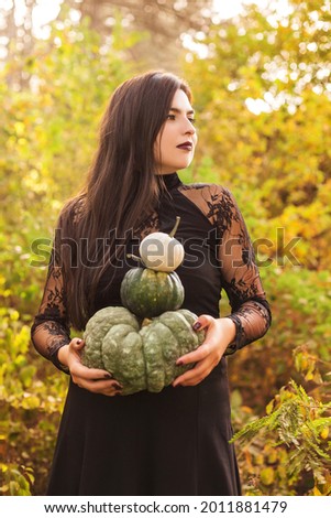Portrait of beautiful young brunette girl with long straight hair and dark vamp makeup looking aside, wearing black guipure dress, holding pumpkins while standing against autumn forest background