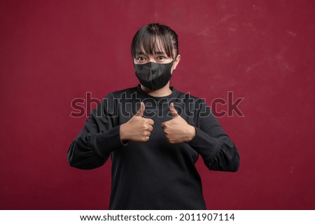 Young beautiful woman wearing black long sleeve t-shirt and protective face mask against corona virus or COVID-19 or filtering dust, air pollution, thumbs showing good sign, on red background