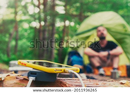 Smartphone is charged using a portable charger. Power Bank charges the phone outdoors with a backpack against the backdrop of a tent and a tourist Royalty-Free Stock Photo #2011936265