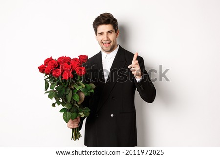 Image of handsome romantic guy in black suit, holding bouquet of roses and pointing at camera, congratulating with holiday, standing against white background