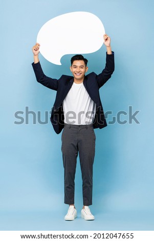 Full body portrait of young happy smiling Asian man holding empty speech bubble standing in light blue isolated studio background Royalty-Free Stock Photo #2012047655