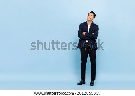 Full length portrait of young handsome southeast Asian businessman with arms crossed looking up to copy space on light blue studio background Royalty-Free Stock Photo #2012065319