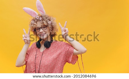 Beautiful girl wearing cute bunny ears hairband and nerdy spectacles making victory signs. Headphones around her neck. Isolated on a pink background studio. Easter concept.