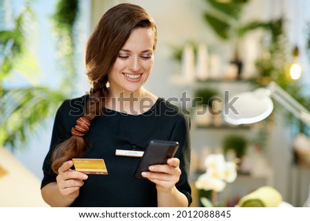 happy female worker with smartphone and credit card making online shopping on e-commerce site in modern beauty salon.