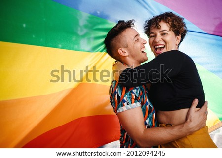 Happy queer couple standing against a rainbow pride flag. Young LGBTQ couple smiling cheerfully while embracing each other. Two non-conforming lovers celebrating gay pride together. Royalty-Free Stock Photo #2012094245