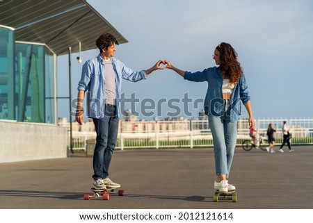Romantic man and woman ride longboard holding hands show heart with fingers. Cute trendy couple in love outdoors enjoy dating relationship activity on skateboards. Urban lifestyle and fashion concept