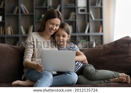 Happy gen Z daughter kid and smiling millennial young mom using laptop on sofa together, shopping on internet, watching movie, online video, enjoying leisure, relaxing with digital device at home