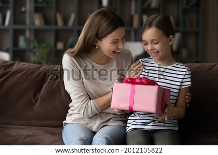 Happy grateful girl giving mothers day gift to mom, holding pink present box. Excited millennial mum and cute daughter kid celebrating birthday, hugging on couch at home, going to unpack festive wrap