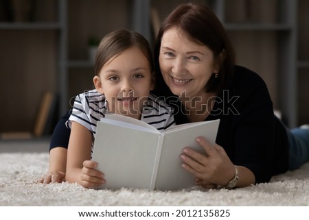 Sweet child reading paper book with granny. Girl and grandmother resting on heating floor at cozy home, looking ant camera, smiling, holding textbook with blank cover. Headshot portrait