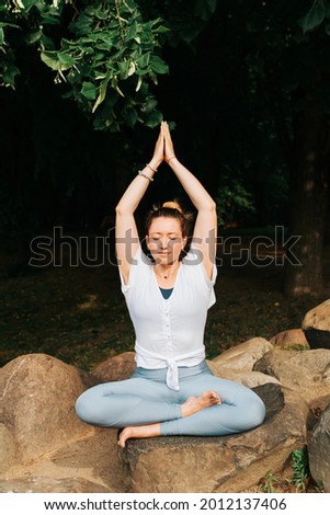Yoga and Meditation. Happy young woman practicing meditation, sitting on a stone in yoga asana in the morning outdoors. Portrait of a smiling yogi woman exercising at sunrise. Vertical photo