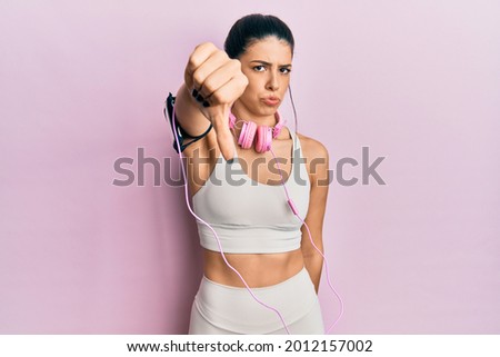 Young hispanic woman wearing gym clothes and using headphones looking unhappy and angry showing rejection and negative with thumbs down gesture. bad expression. 