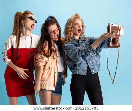 Vintage selfie. Pretty young women in retro 90s fashion style, outfits isolated over blue studio background. Concept of eras comparison, beauty, fashion and youth. Look happy, excited, delighted Royalty-Free Stock Photo #2012165783