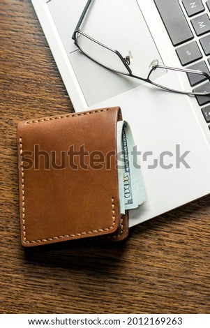 Brown leather wallet with money ($100 USD), glasses and laptop keyboard on wooden table. Internet payments, online shopping, startup business profit concept, copy space. Flat lay, top view