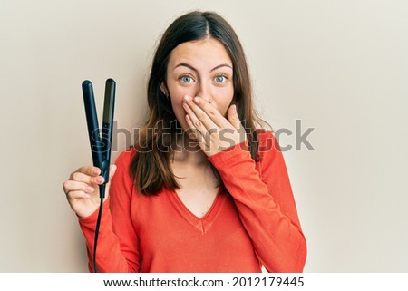 Young brunette woman holding hair straightener covering mouth with hand, shocked and afraid for mistake. surprised expression 