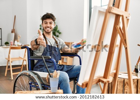 Young hispanic man sitting on wheelchair painting at art studio smiling cheerful offering palm hand giving assistance and acceptance. 