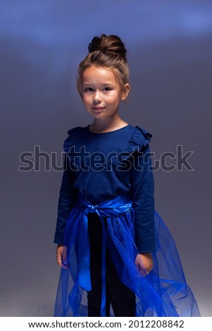 A fashionable portrait of a little model girl posing professionally on a dark background. A girl in black leggings, a blue jacket and a blue train. The concept of children's fashion