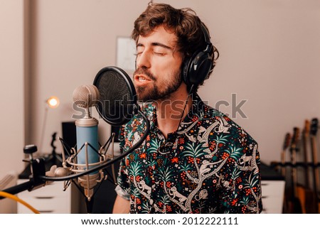 Millenial male singer on headphones singing into microphone while working on songs during a recording studio session at home.