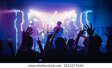 Rock Band with Guitarists and Drummer Performing at a Concert in a Night Club. Front Row Crowd is Partying. Silhouettes of Fans Raise Hands in Front of Bright Colorful Strobing Lights on Stage. Royalty-Free Stock Photo #2012271626