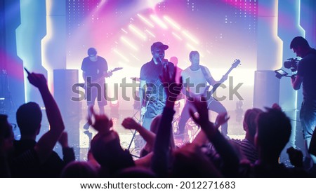 Rock Band with Guitarists and Drummer Performing at a Concert in a Night Club. Front Row Crowd is Partying. Silhouettes of Fans Raise Hands in Front of Bright Colorful Strobing Lights on Stage. Royalty-Free Stock Photo #2012271683