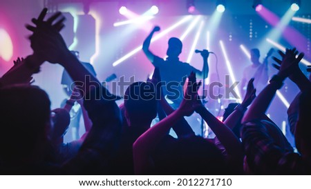 Rock Band with Guitarists and Drummer Performing at a Concert in a Night Club. Front Row Crowd is Partying. Silhouettes of Fans Raise Hands in Front of Bright Colorful Strobing Lights on Stage. Royalty-Free Stock Photo #2012271710