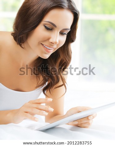 Image of beautiful happy smiling amazed woman using tablet pc, touchpad, ipad looking at, in bed room, near window. Attractive brunette girl at home bedroom. Online, distance, zoom.