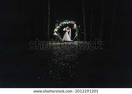 Stylish groom in a vest and a cute brunette bride in a white long dress with a veil are dancing, hugging near a wooden arch, decorated with flowers, lamps, in the forest at night. Wedding photography.