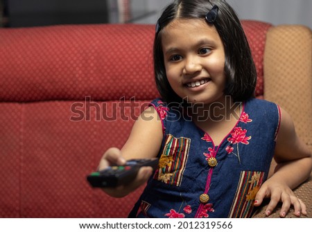An Indian girl child smiles while watching television with remote in hand and sitting on sofa