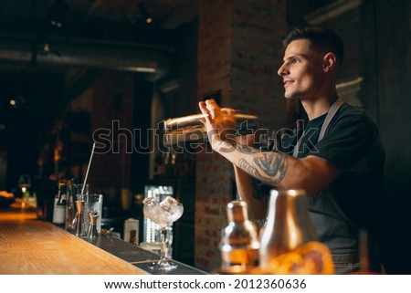 Young handsome smiling male bartender in apron preparing alcohol drink shaking it in cocktail shaker standing in front of bar counter. Side view Royalty-Free Stock Photo #2012360636
