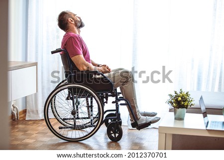 Handicapped person sitting in wheelchair and thinking about life, depression. Disabled mature man sitting on wheelchair and looking out the window in the living room at home