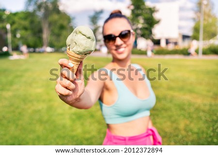 Fit slim woman showing ice cream cone in city park in summer. Happy smiling portrait of young laughing model and melting icecream. Stylish trendy bikini and sunglasses. Scoop of gelato. Outdoor joy.