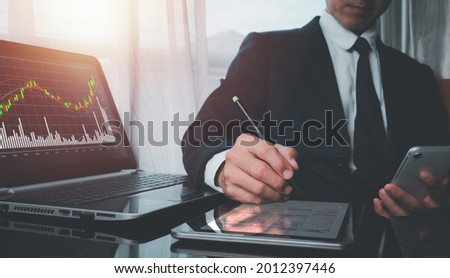 A man wearing a suit on his left hand holding a cell phone His right hand poked a pen on a tablet's screen, and the screen of the laptop shows the fast-growing stock chart.