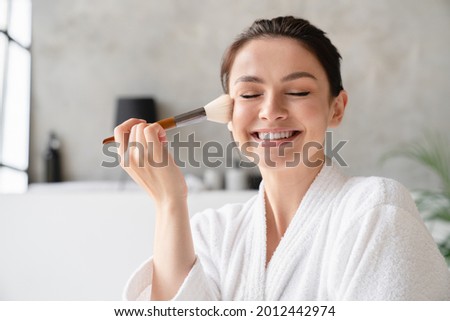 Smiling happy young woman applying makeup on her face with cosmetics brush in white spa bathrobe making morning preparations. Beauty care and treatment Royalty-Free Stock Photo #2012442974