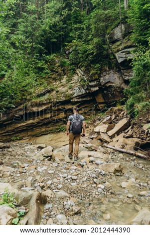 Guy tourist on a background of large mountain stones and coniferous trees. A young man with a tourist backpack crosses the rocks of a mountain river.