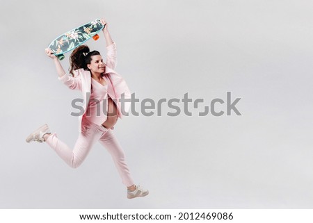 A pregnant girl in a pink suit with a skateboard in her hands jumps on a gray background
