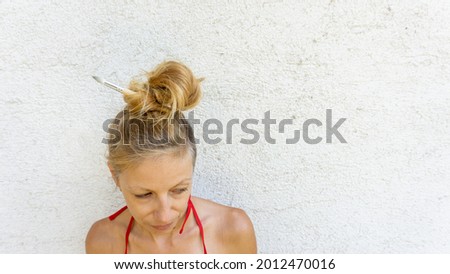 Beautiful blond female person wiht white wall in the background showing emotions and loking in different directions while having paintbrushes in the hair. Single stylish female model.  