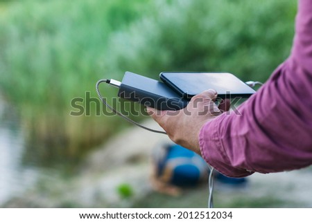 The guy is holding a portable charger with a smartphone in his hand. Man on a background of nature with a greenery and a lake.