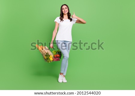 Full size photo of funny brunette young lady hold food show thumb up wear t-shirt jeans isolated on green color background