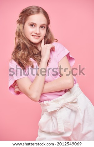 Cute teenage girl with blonde hair and in a pink t-shirt smiles at camera on a pink background. Positive emotions. 