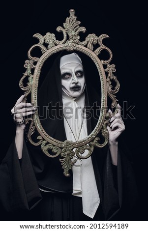 Halloween. Portrait of a scary devilish nun with a picture frame in her hands on a black background. Horrors.