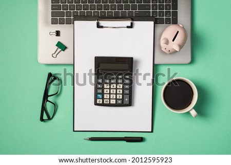 Top view photo of green and black binder clips piggy bank on grey laptop glasses cup of drink pen and calculator on black clipboard folder with paper sheet on isolated turquoise background