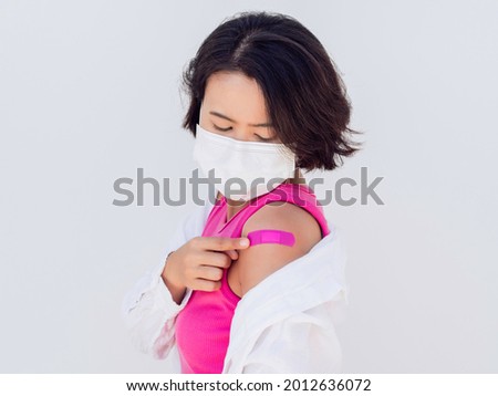 Vaccinations concept. Vaccinated Asian woman wearing a face mask, pink sleeveless and white shirt looking at pink bandage plaster on her shoulder after vaccination treatment isolated on white.