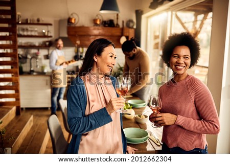 Happy African American woman and her Asian female friend drinking wine while their husbands are preparing dinner in the background. 