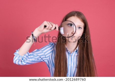 Little kid girl 13 years old in blue dress isolated on pink background does grimaced in magnifier