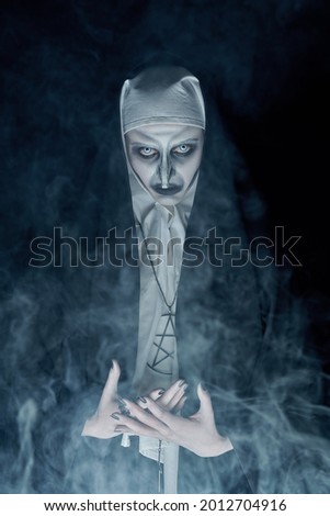Halloween and horrors. Portrait of a scary devilish nun looking at camera on a black background with mysterious smoke. 
