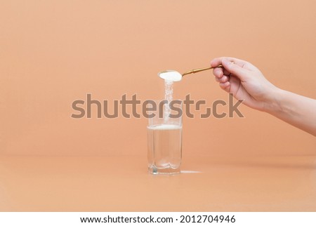 Hand pours collagen protein powder in a glass of water on a beige background. A natural supplement for skin beauty and bone health. Space for text. Royalty-Free Stock Photo #2012704946