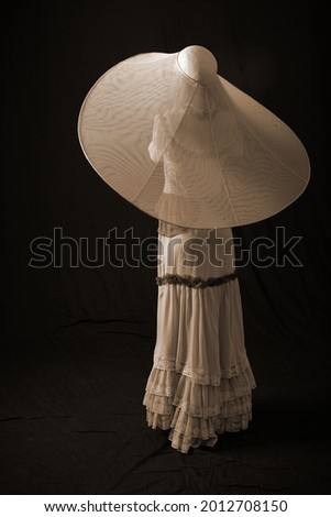 A girl in an old dress and hat stands on a dark background and is sad