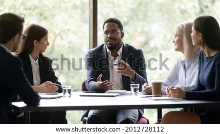 Engaged motivated African American business leader talking to team, meeting with employees, managers. Corporate coach, mentor training staff. Diverse group discussing work project at table Royalty-Free Stock Photo #2012728112
